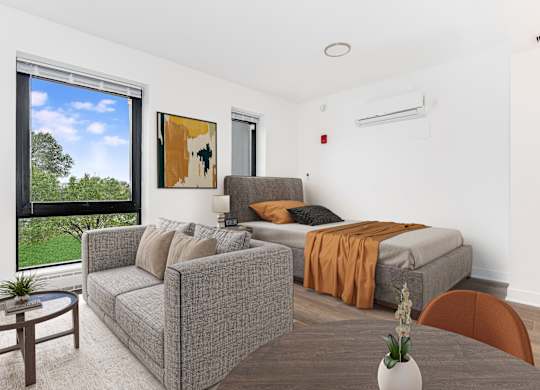 A living room with a grey couch, a brown chair, a bed with an orange blanket, and a painting on the wall.