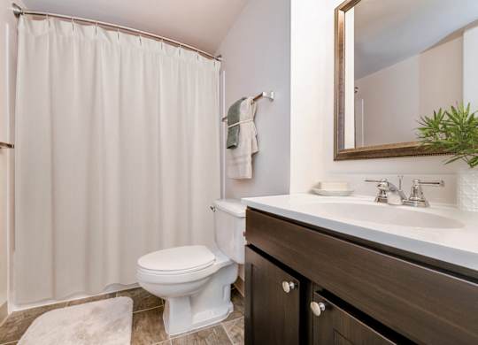 A white toilet sits in a bathroom with a white shower curtain and a mirror above a sink.