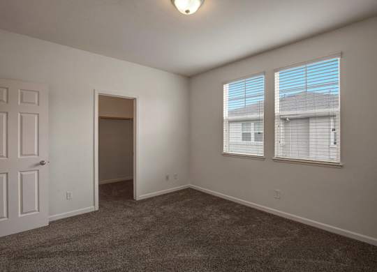 A room with a carpeted floor, two windows with blinds, and a door.