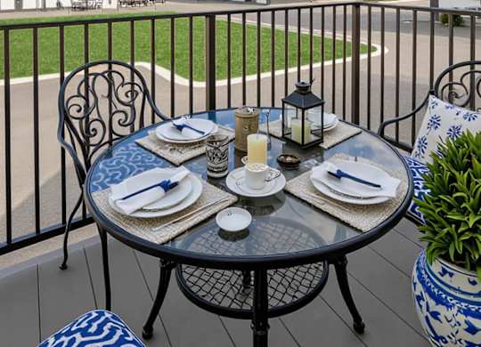 A table set for a meal on a balcony with a view of a white house and a blue sky.