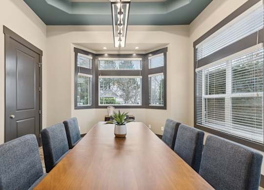 A long brown table is surrounded by grey chairs in a room with a large window.