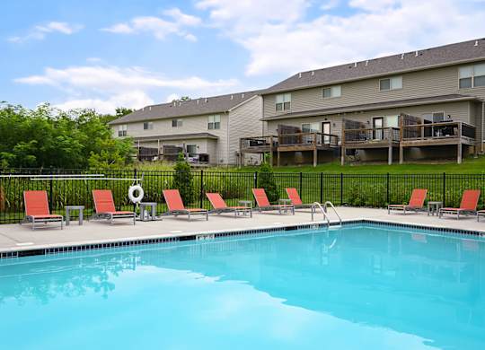 A swimming pool in front of a building with red chairs around it.