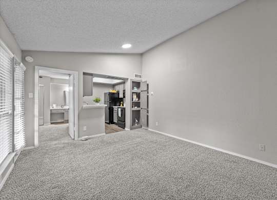 A spacious room with a carpeted floor and a kitchen area visible through the open door.