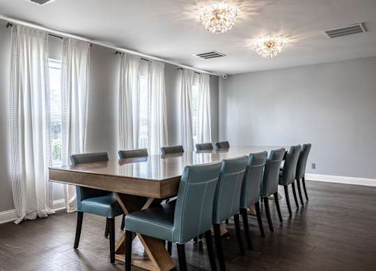 A long wooden table with blue chairs is in the middle of a room with a window.