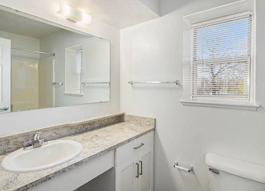A bathroom with a sink, mirror, toilet, and a window with blinds at Black Sand Apartment Homes in Lincoln, NE