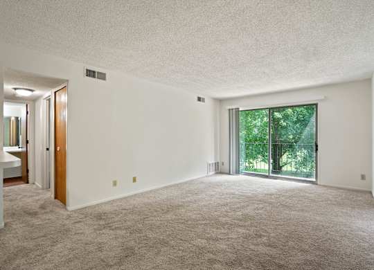 A large, empty room with a carpeted floor and a sliding glass door leading to a green outdoor area at Beacon Hill and Great Oaks Apartments, Rockford, Illinois
