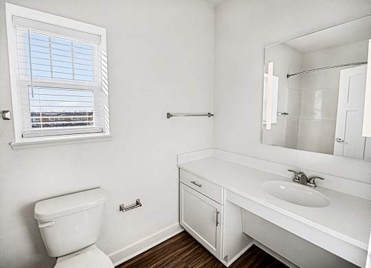 A white bathroom with a toilet, sink, mirror and a window at The Grove Apartment Homes in Lincoln, NE