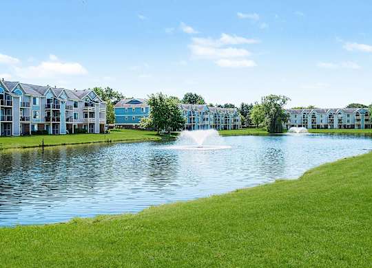 A serene pond with a fountain in the middle of a grassy area with buildings in the background at Indian Lakes Apartments, Mishawaka, Indiana