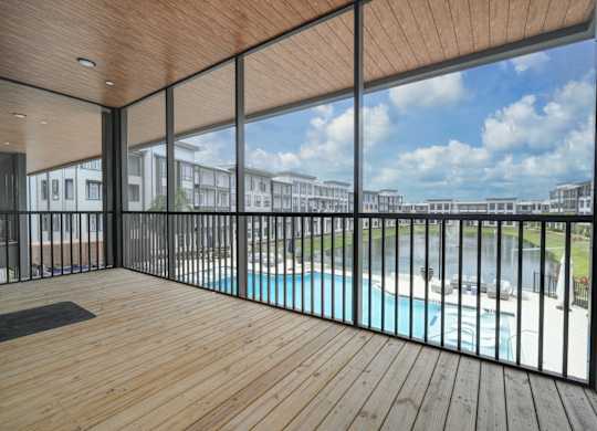 A wooden deck with a glass railing overlooking a pool and buildings.