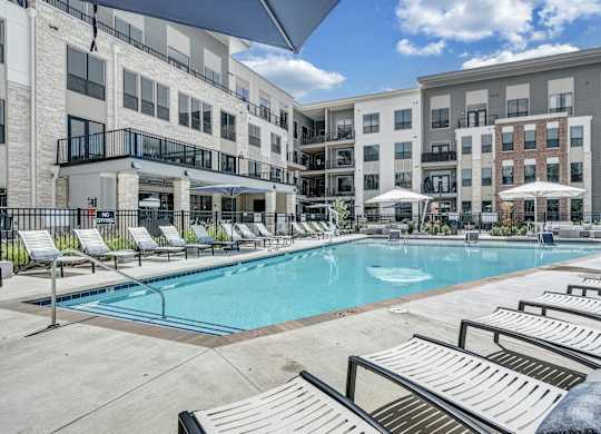 A swimming pool surrounded by sun loungers and umbrellas in front of apartment buildings at Luxe 360 Apartment Complex in Midlothian Virginia