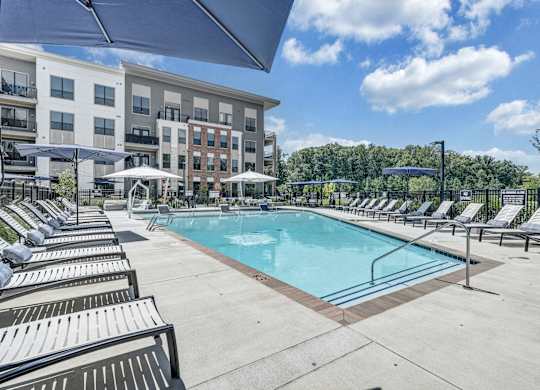 A swimming pool surrounded by sun loungers and umbrellas in front of apartment buildings at Luxe 360 Apartment Complex in Midlothian Virginia