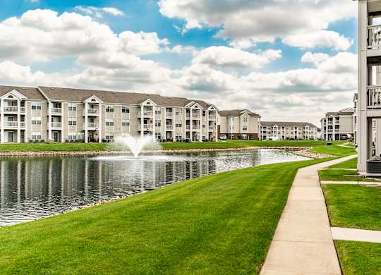 A row of apartment buildings are lined up next to a pond with a fountain in the middle at Mallard Bay Apartments, Crown Point, Indiana