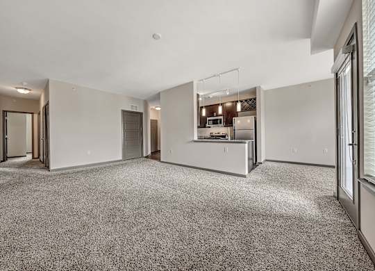 A spacious, empty room with a carpeted floor and a kitchen area in the background.at River Crossing Apartments, St. Charles, MO