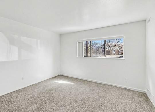 A garden style living room with a carpeted floor and a window showing a view of trees outside at Trappers Cove Apartments, Lansing