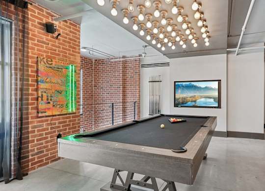 A pool table is in the middle of a room with a brick wall and large TV