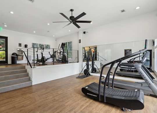 A gym with a treadmills, equipment, a staircase, and a ceiling fan.