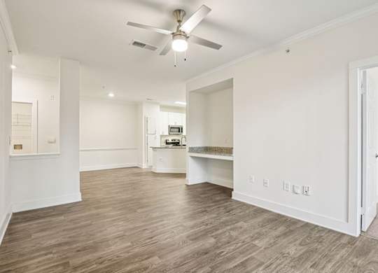 A spacious apartment . A view of the living room, dining room, and kitchen. There's a ceiling fan and light fixture, hardwood floors, and white walls with a doorway leading to a bedroom.