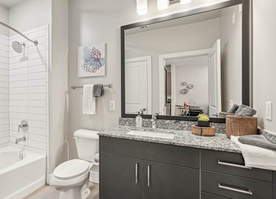 A bathroom with a white tub and shower, toilet, and granite countertop with a sink.