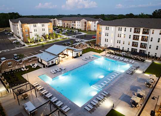 A large swimming pool surrounded by lounge chairs and umbrellas in front of apartment buildings.