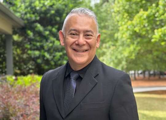 A man in a black suit and tie standing in front of a greenery.