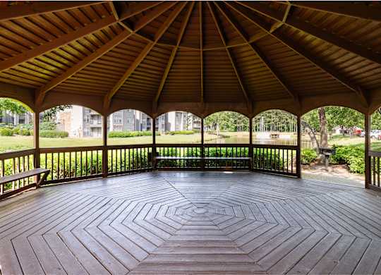 A wooden pavilion with a circular floor and a roof next to a pond and grill