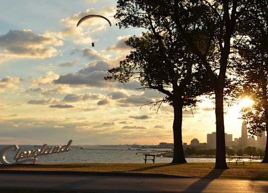 A person is paragliding over a park with trees and a Cleveland sign in the background.