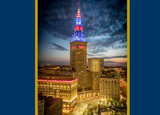 High-angle twilight view of The Terminal Tower Residences illuminated in patriotic red, white, and blue lights, soaring above the cityscape of downtown Cleveland.