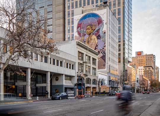 A tall building with a blue top stands next to a building with a mural on it.