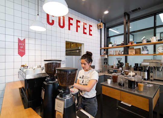 A woman making coffee with a white wall behind her that says coffee.