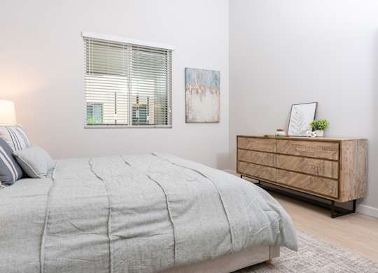 A bed with a grey comforter and pillows sits in a room with a wooden dresser and a painting on the wall.