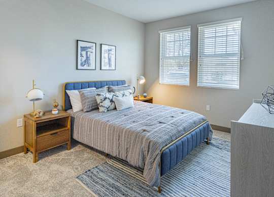 Studio Apartments In Burlingame, CA - Bayswater Burlingame - Spacious Bedroom With Plush Carpet Flooring, Area Rug, Bed, Nightstands With Lamps, Dresser, Wall Art, And Two Windows With Blinds at Baysw