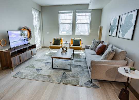 1-Bedroom Luxury Apartments In Burlingame, CA - Bayswater Burlingame - A Modern Living Room With A Grey Sofa, Yellow Accent Chairs, A Geometric Rug, And A Large Flat-Screen Television.