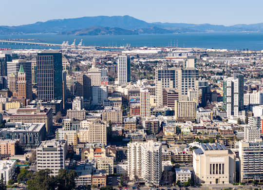 A cityscape with a mix of modern and older buildings, a bridge in the distance, and a mountain range in the far background.
