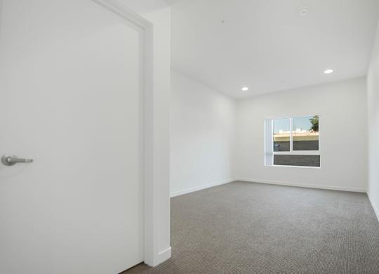 A white door is on the left of a room with a carpeted floor and a window with a view of a building.
