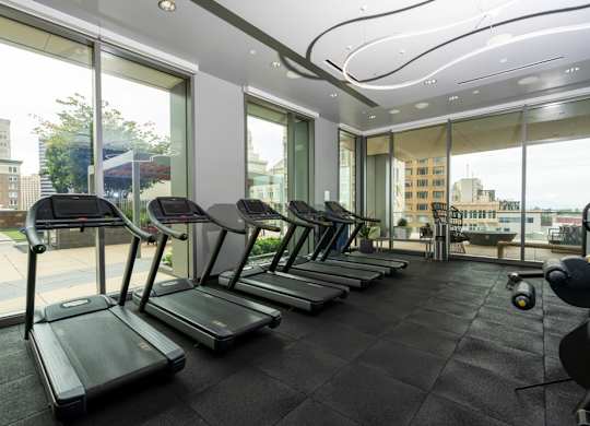 A row of treadmills are lined up in a gym with a view of the city outside the windows.