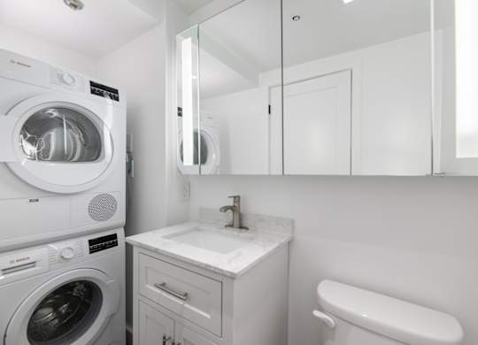 A white washing machine is in a small laundry room with a sink and mirror.