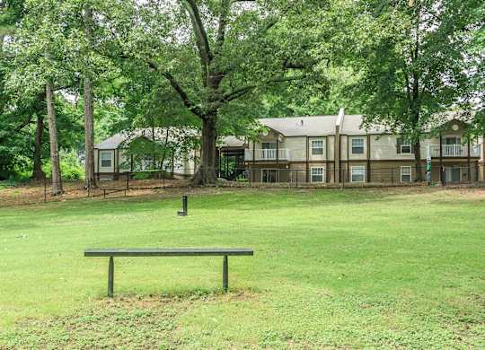 A bench sits in the middle of a grassy field with apartment buildings in the background.