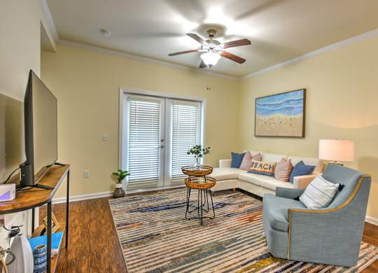 A living room with a grey sofa, a wooden table, a television, a ceiling fan, and a painting of a beach scene on the wall at Cabana Club - Galleria Club Apartments, Jacksonville, 32256