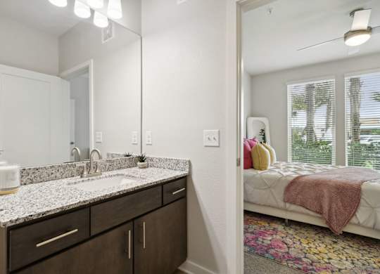 A bathroom with a sink, mirror, and a bedroom with a bed and windowat Pinnacle Apartments, Jacksonville, FL