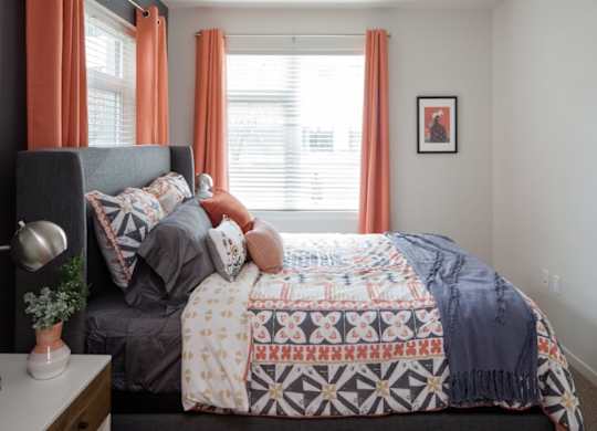 A bed with a patterned comforter and a grey headboard with a lamp and a plant on the nightstand.