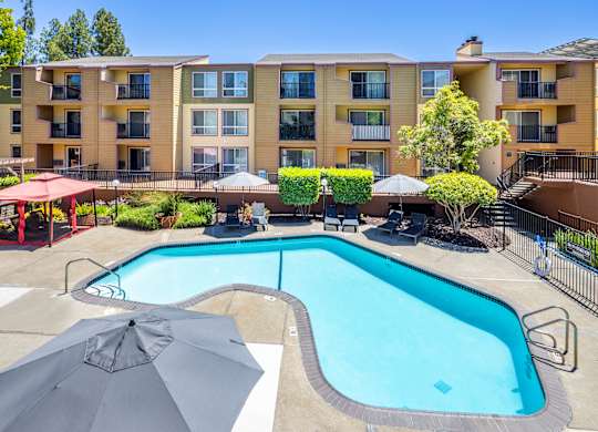 A large swimming pool surrounded by a fence and umbrellas in front of a multi-story apartment building.