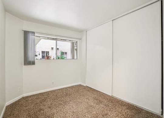 A room with a carpeted floor and a window showing a view of a house across the street.