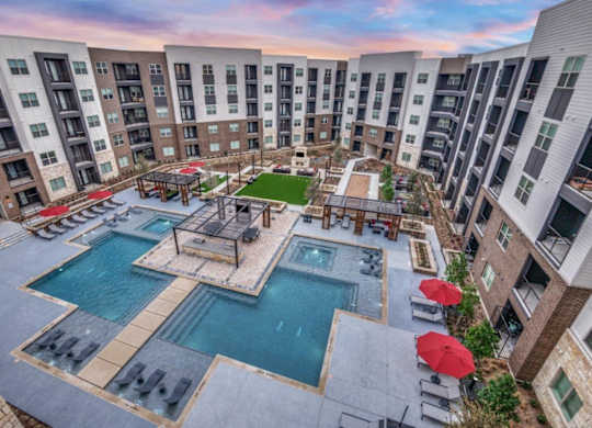 A large swimming pool surrounded by red umbrellas and lounge chairs in a courtyard surrounded by apartment buildings.