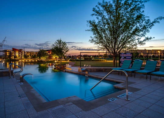 A pool surrounded by chairs and a tree at dusk.