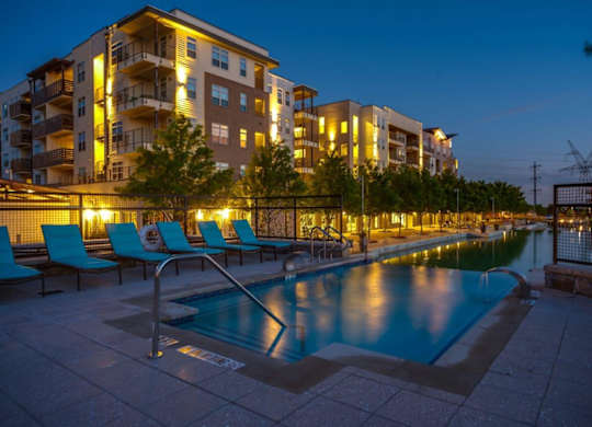 A swimming pool is surrounded by chairs and is lit up at night.