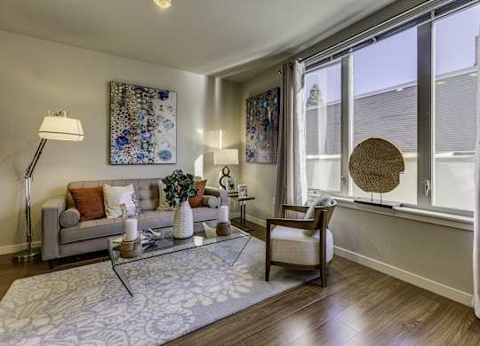 A living room with a grey couch, a glass coffee table, and a large window with a wicker chair.