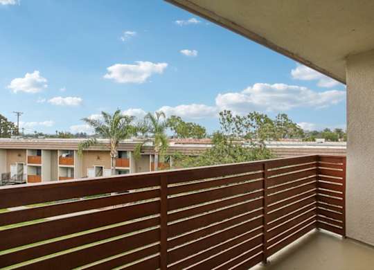 A balcony with a brown railing overlooks apartment buildings and trees.