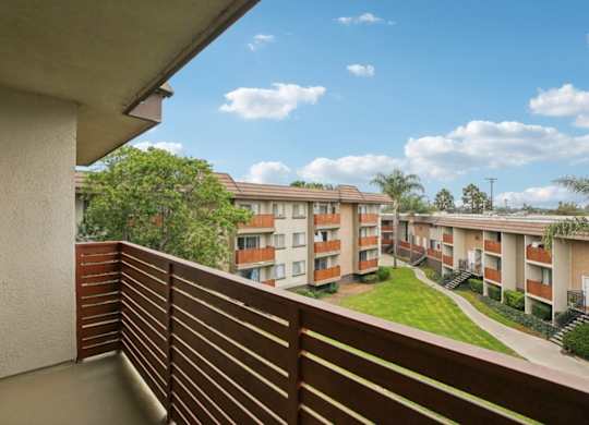 A balcony with a wooden railing overlooks a grassy courtyard and apartment buildings.