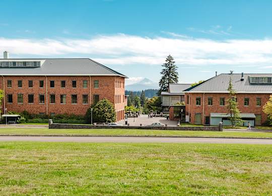 A large red brick building with a black roof is surrounded by a green field.