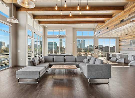 A large grey couch sits in the middle of a spacious room with wooden floors and a city view through the windows.
